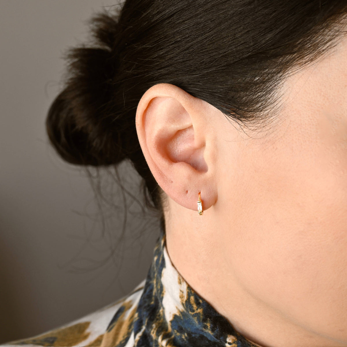 Gold-tone huggie earring with a rectangular clear stone worn on an earlobe.