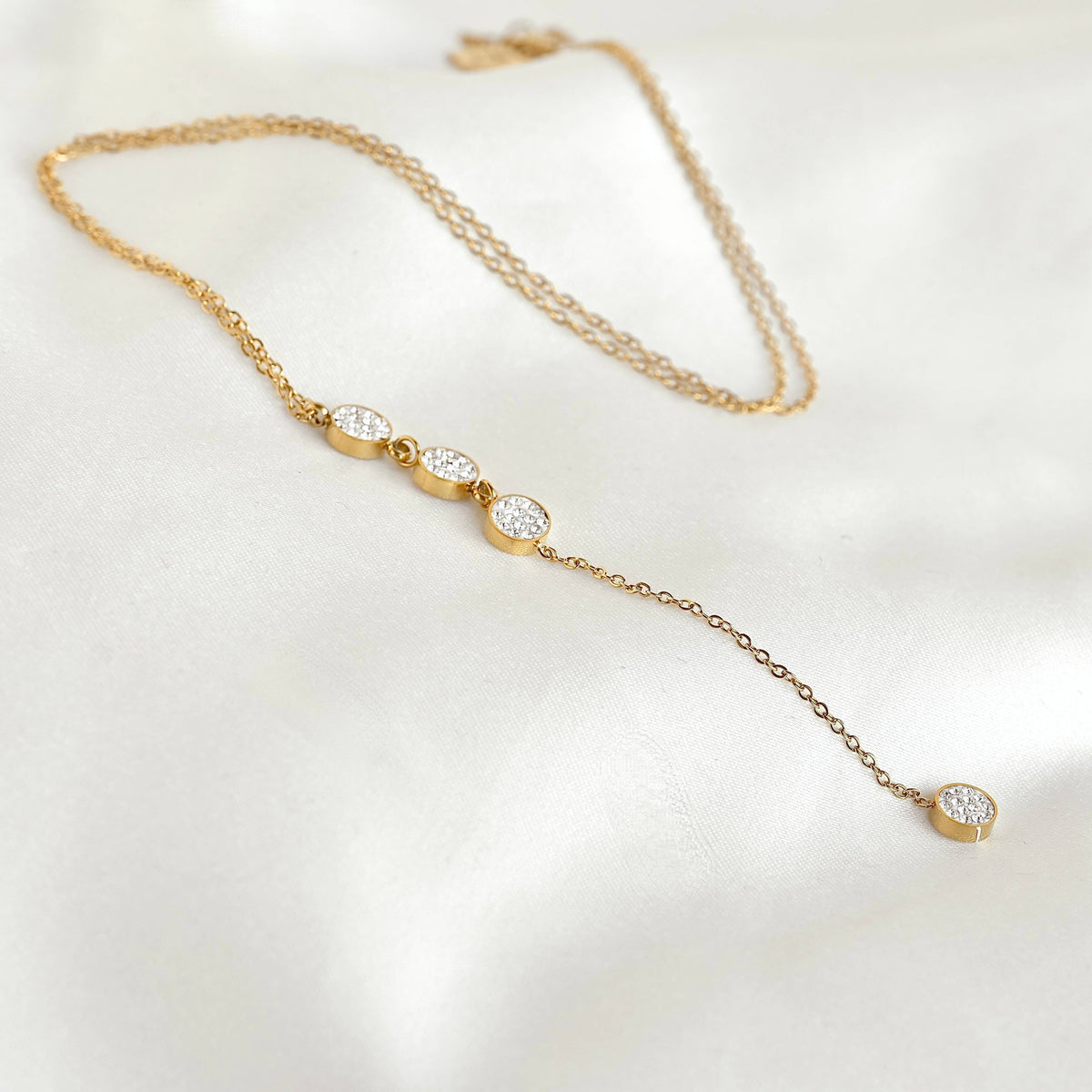 Gold pavé circle lariat necklace arranged on white fabric, showing the fine chain, three linked crystal circles and long pendant drop.