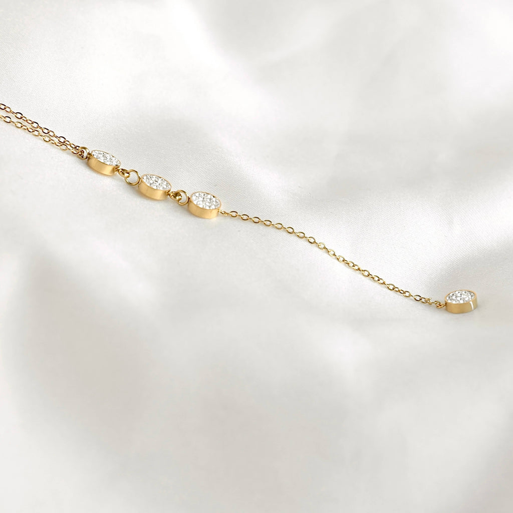 Close-up of a gold lariat necklace on white fabric, showing pavé crystal circles and the chain leading to the final round drop.