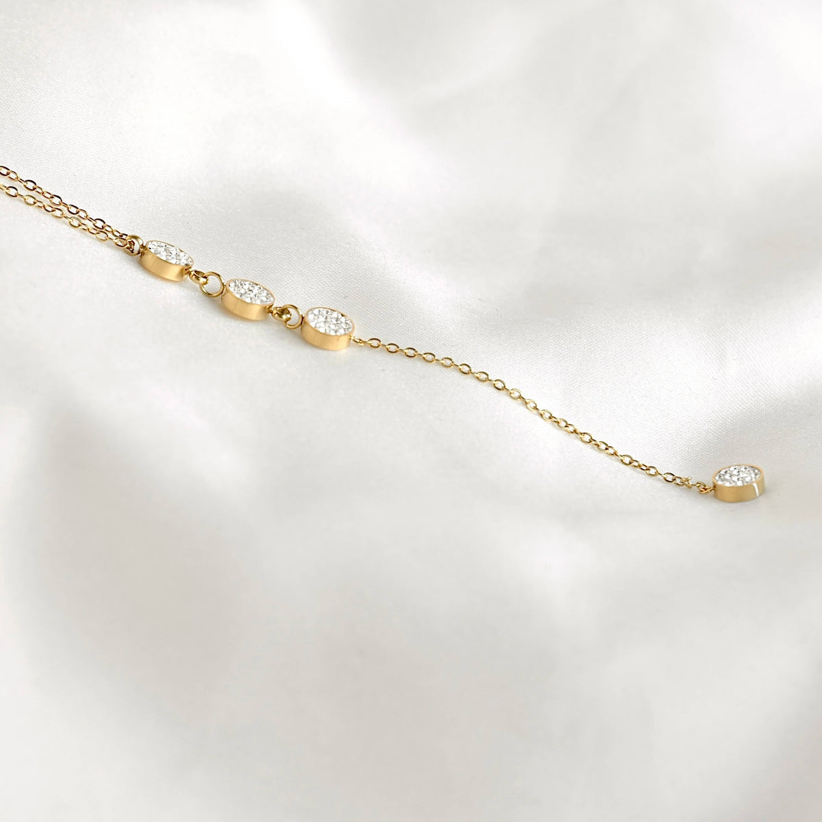 Close-up of a gold lariat necklace on white fabric, showing pavé crystal circles and the chain leading to the final round drop.