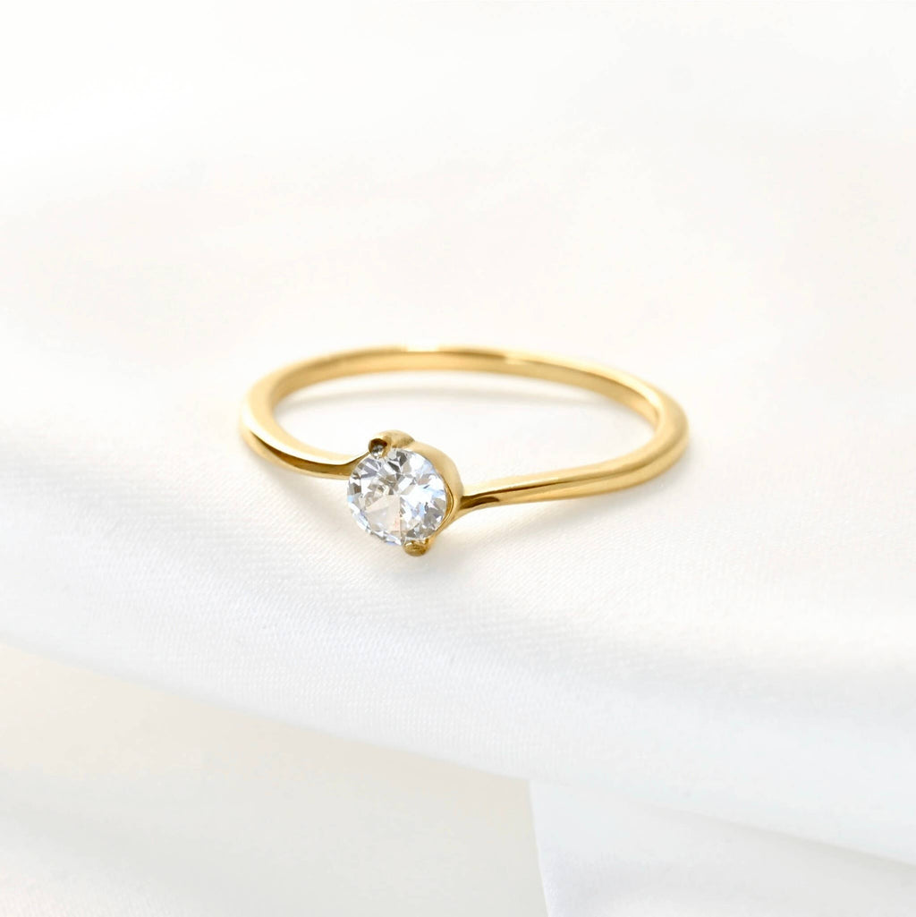 Gold bypass solitaire ring lying on a white surface, showing the slender band curving around a round clear stone.
