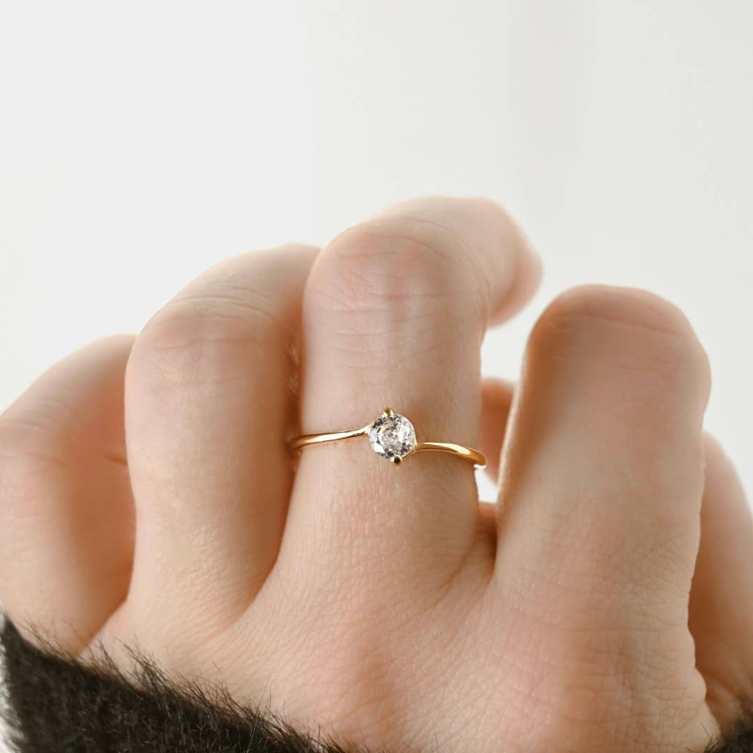 Gold bypass solitaire ring worn on a hand beside a dark knit cuff, showing the delicate band and single clear stone.