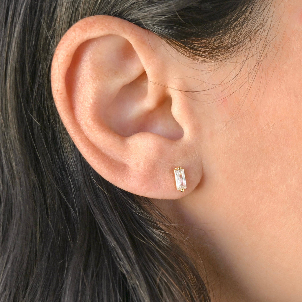 Close-up of a clear bar stud in a gold-tone setting worn alone on an earlobe.