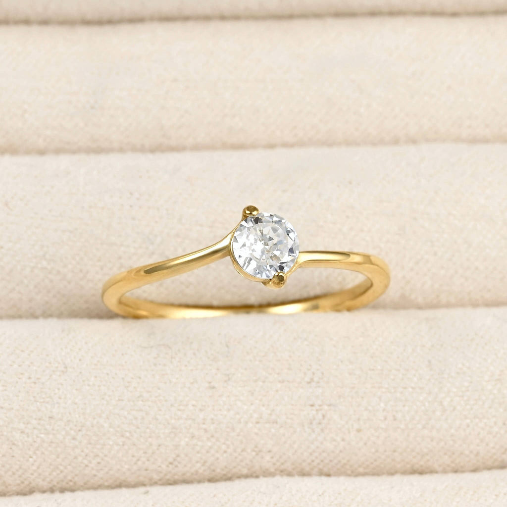 Close-up of a gold bypass solitaire ring with a round clear stone and curved polished band in a cream jewellery tray.