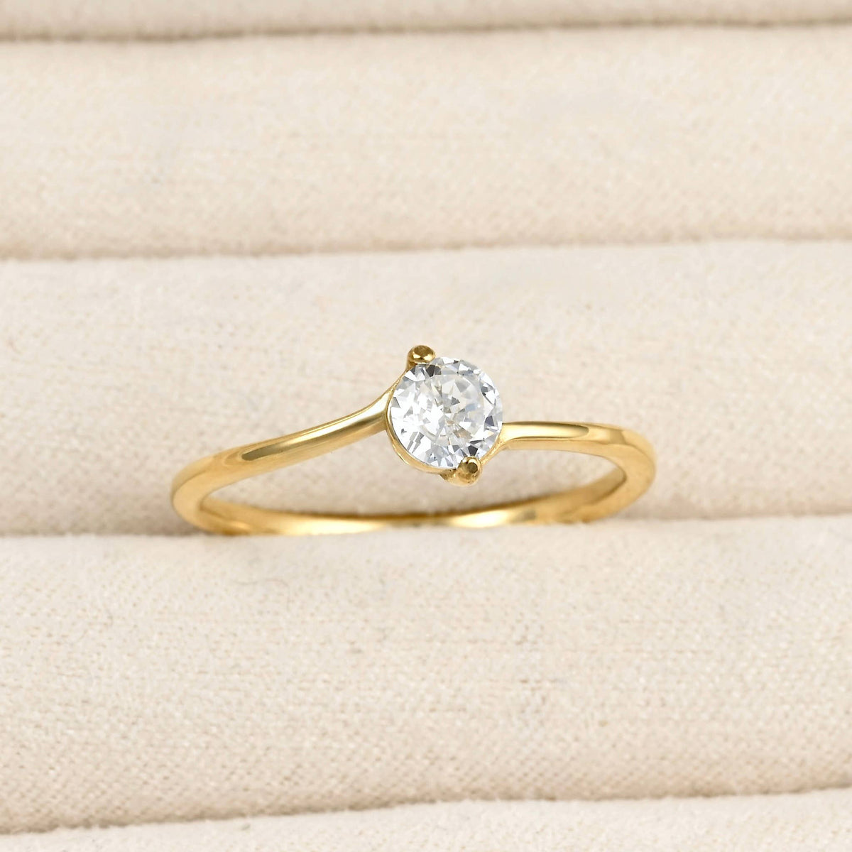 Close-up of a gold bypass solitaire ring with a round clear stone and curved polished band in a cream jewellery tray.