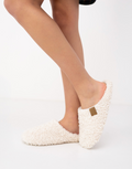 "Cream Sensis Nomi slippers with a plush textured finish and a small tan label patch, worn by a model against a white background." 