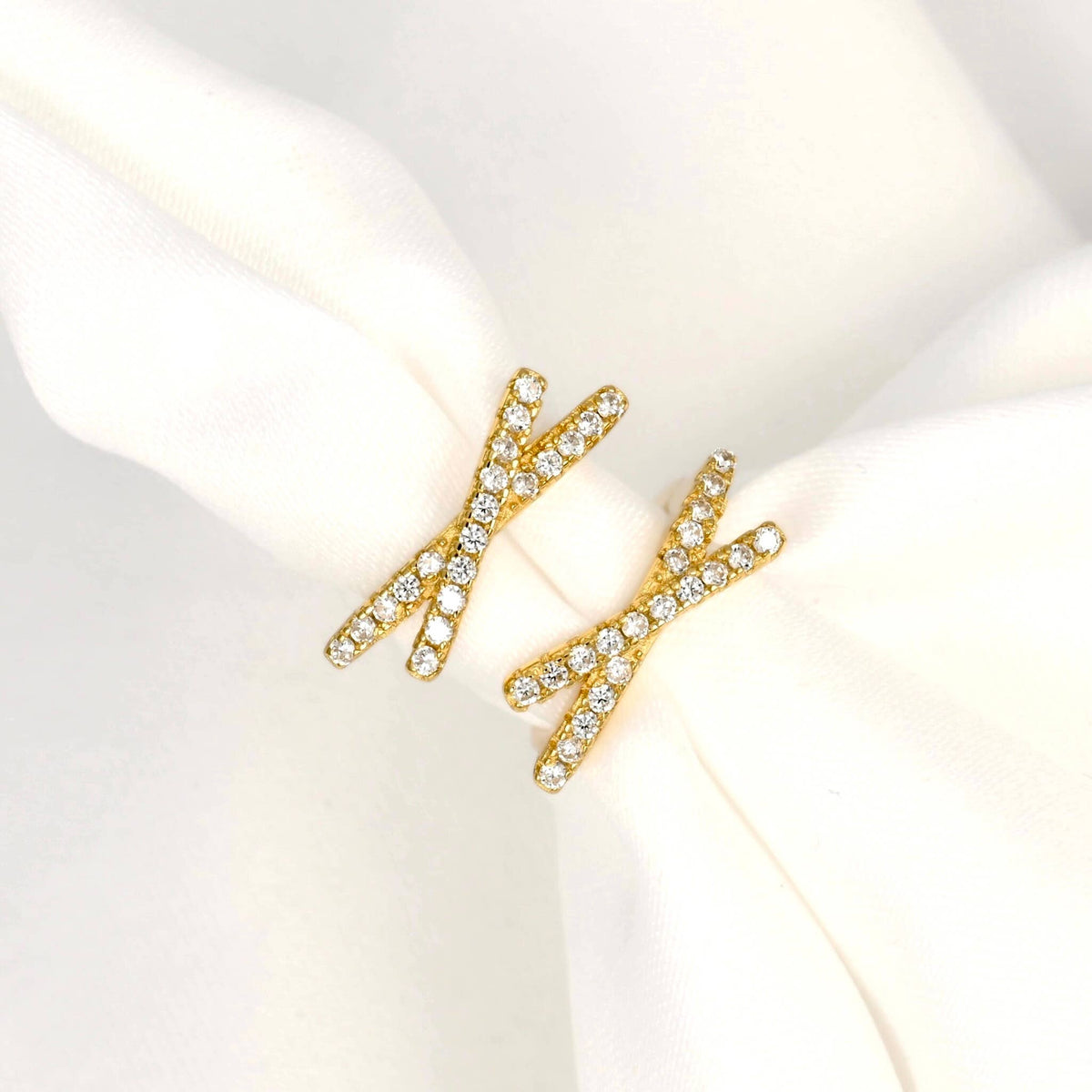 Front view of gold-tone criss cross hoop earrings on white fabric, highlighting the clear stones and X-shaped design.