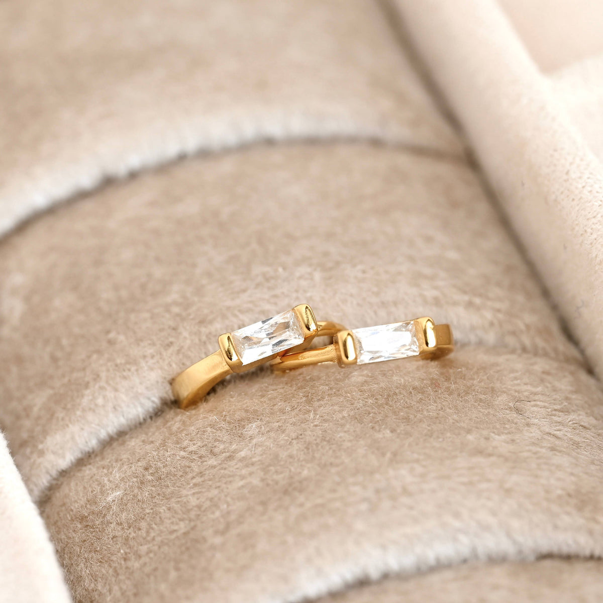 Gold-tone huggie earrings with rectangular clear stones displayed in a cream jewellery tray.