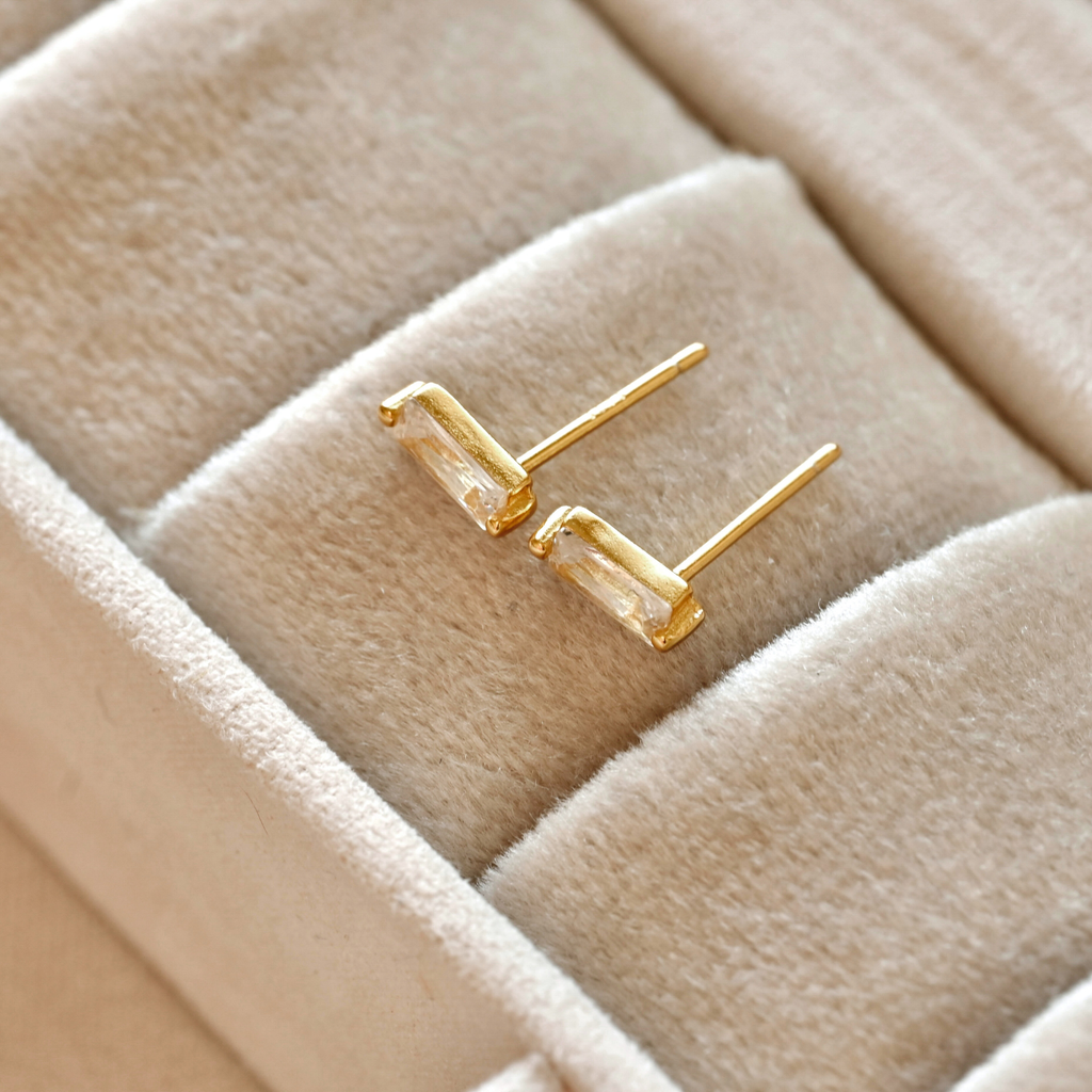 Side view of gold-tone rectangular bar studs in a cream jewellery tray, showing clear stones and straight posts.