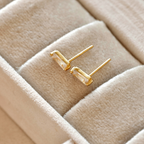 Side view of gold-tone rectangular bar studs in a cream jewellery tray, showing clear stones and straight posts.