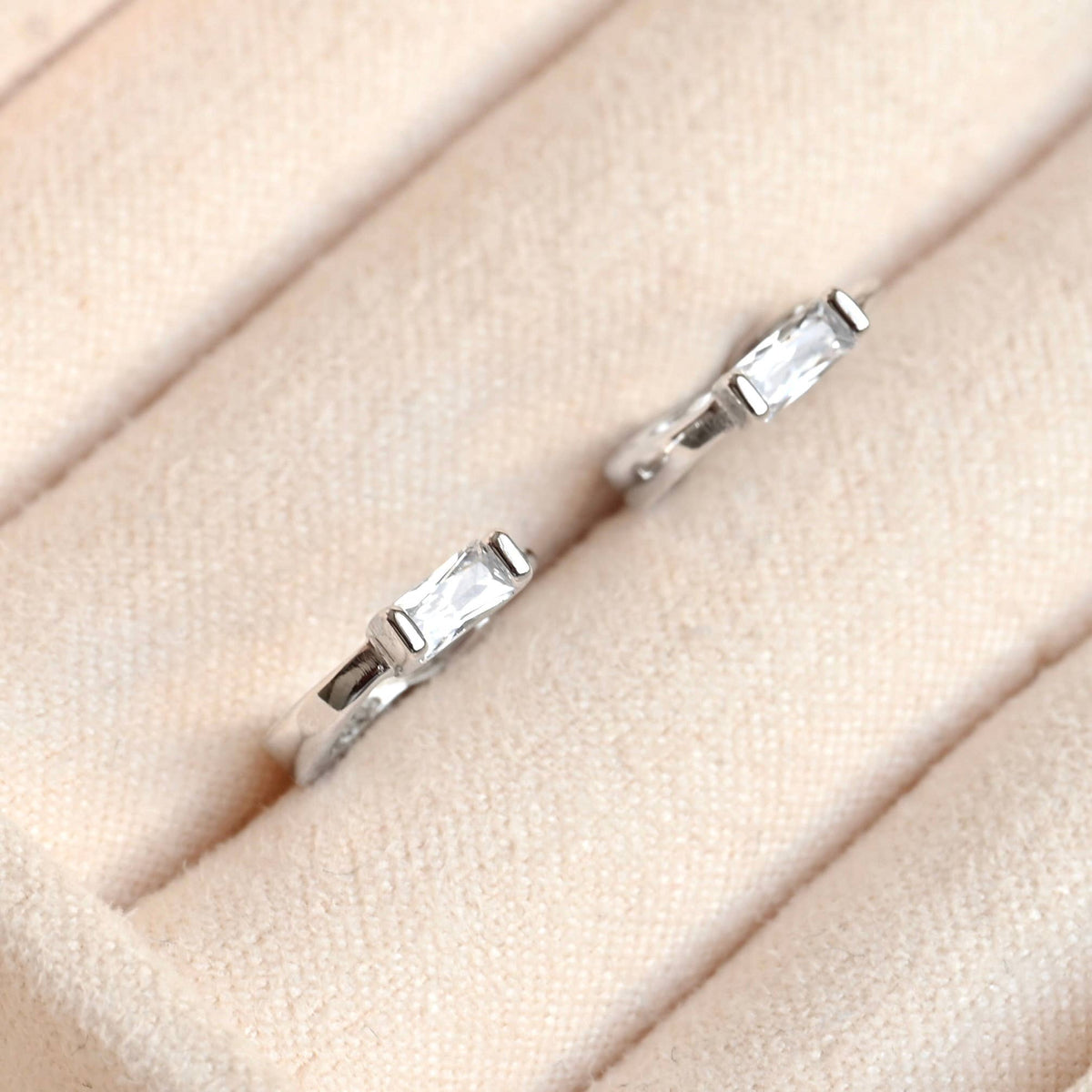 Pair of silver-tone huggie earrings with rectangular clear stones in a cream jewellery tray.