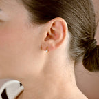 Gold-tone huggie hoop with a round clear stone worn on an earlobe, with hair swept back.
