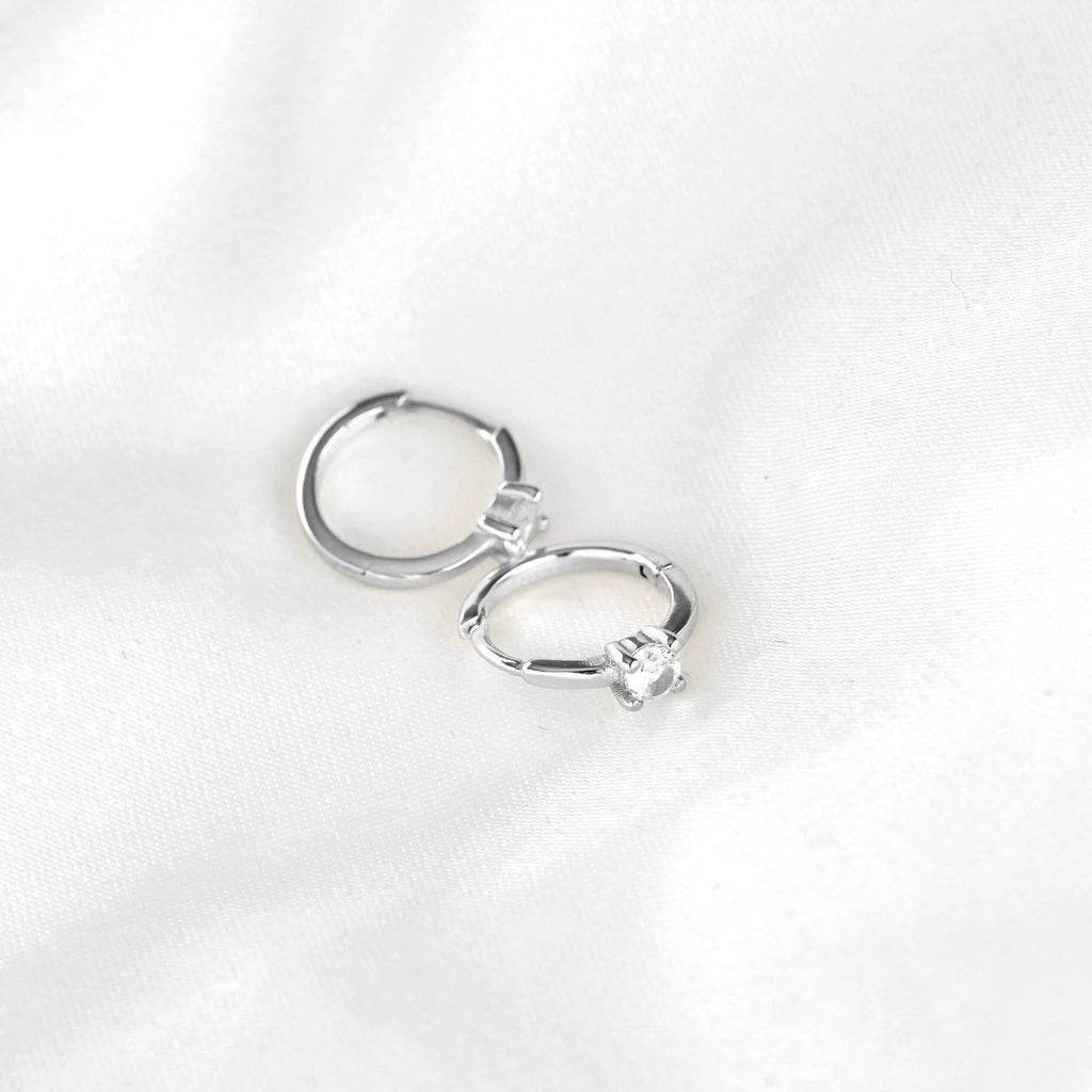 Silver-tone huggie hoop earrings with round clear stones resting on white fabric, showing hinged closures.