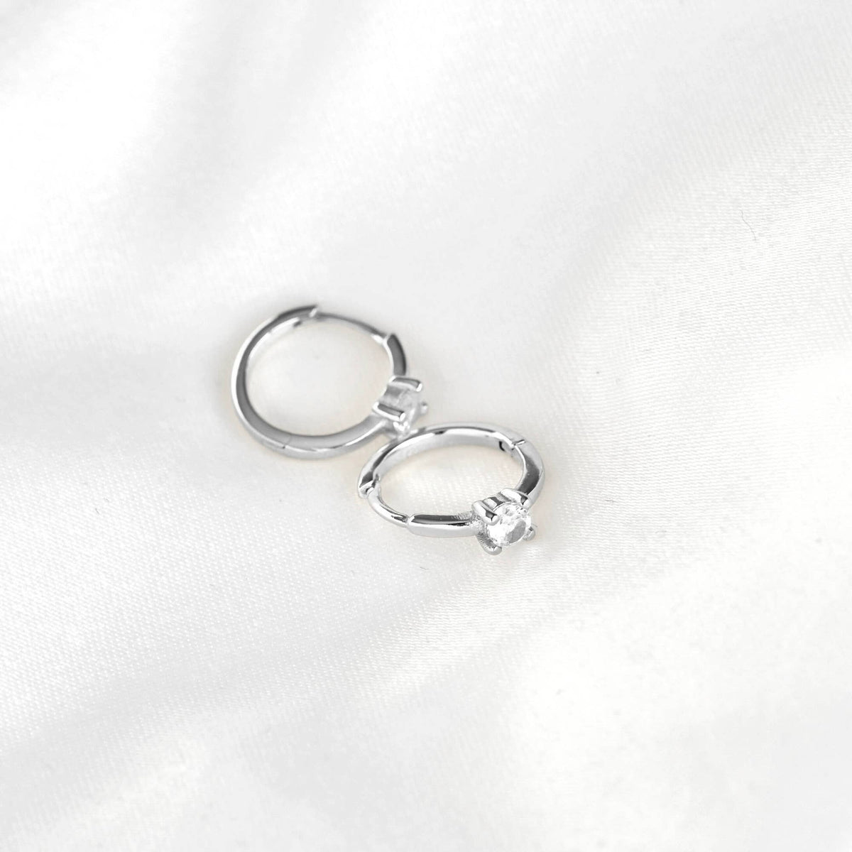 Silver-tone huggie hoop earrings with round clear stones resting on white fabric, showing hinged closures.